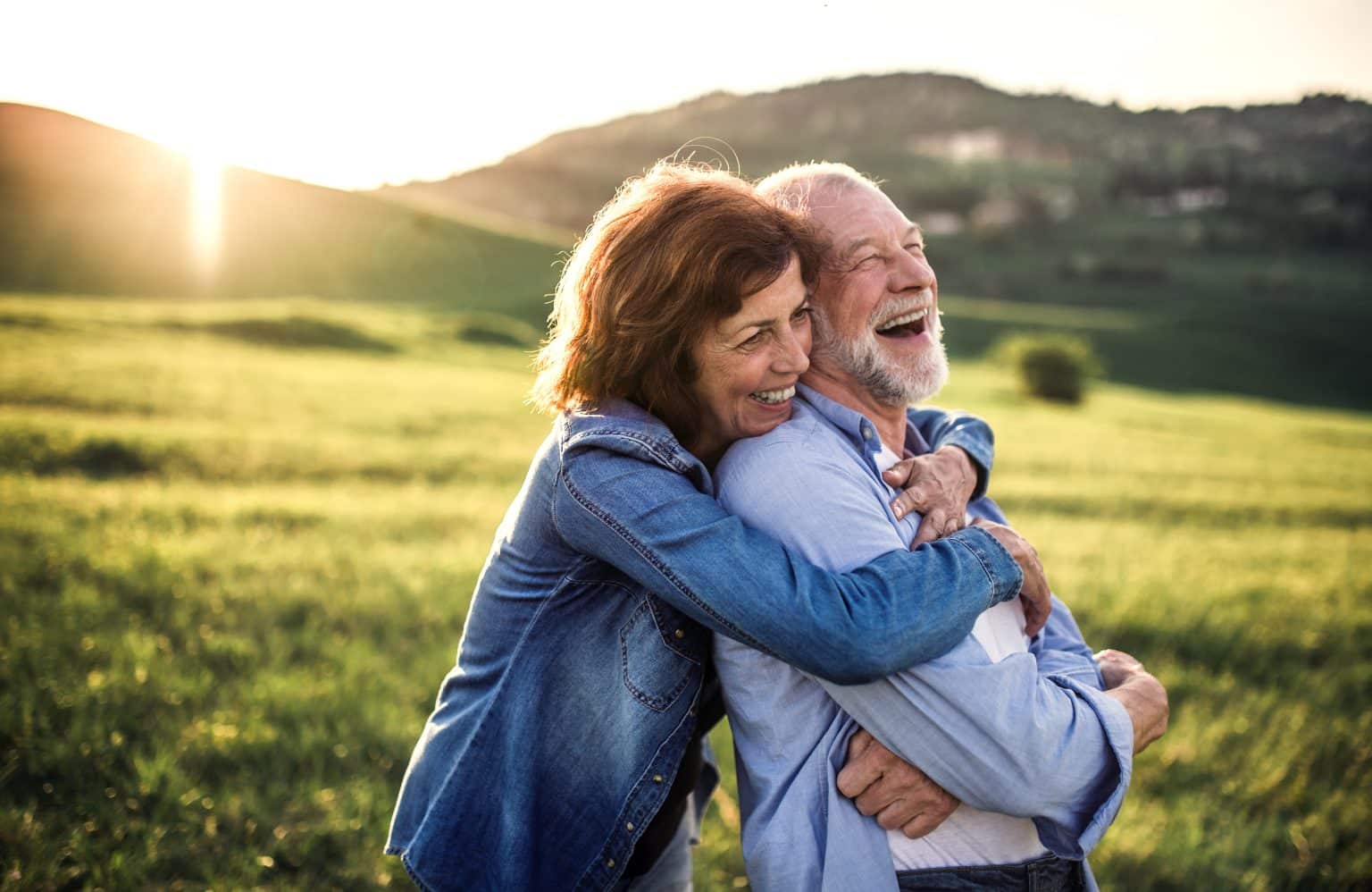 Woman holds a man in a field from behind. He is leaning into her and laughing with his arms crossed. Her arms are draped over his shoulders. She is smiling and the sun is going down on the hillside behind them. 