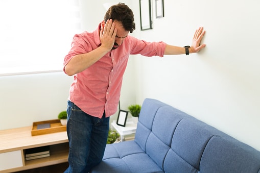 Man leaning against wall in his living room, holding his head with one hand while his other is outstretched against the wall for support