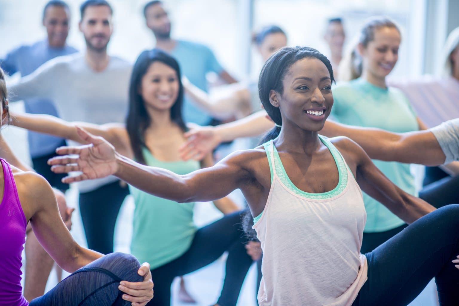 Woman smiling in an exercise class with arm outstretched. She is balancing in a group class with men and woman. She is holding her knee with her hand. 